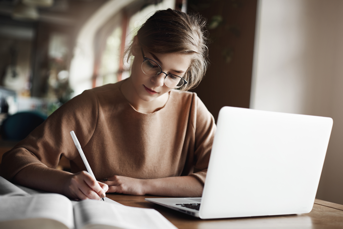 hardworking-focused-woman-trendy-glasses-concentrating-writing-essay-sitting-cozy-cafe-near-laptop-working-making-notes-carefully